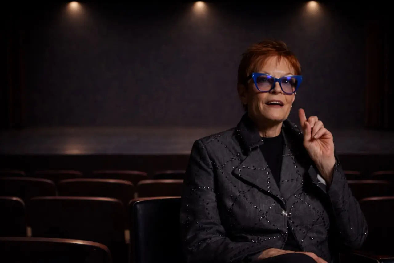 A woman with short red hair and blue glasses sits in an empty theater, gesturing with her right hand while speaking.