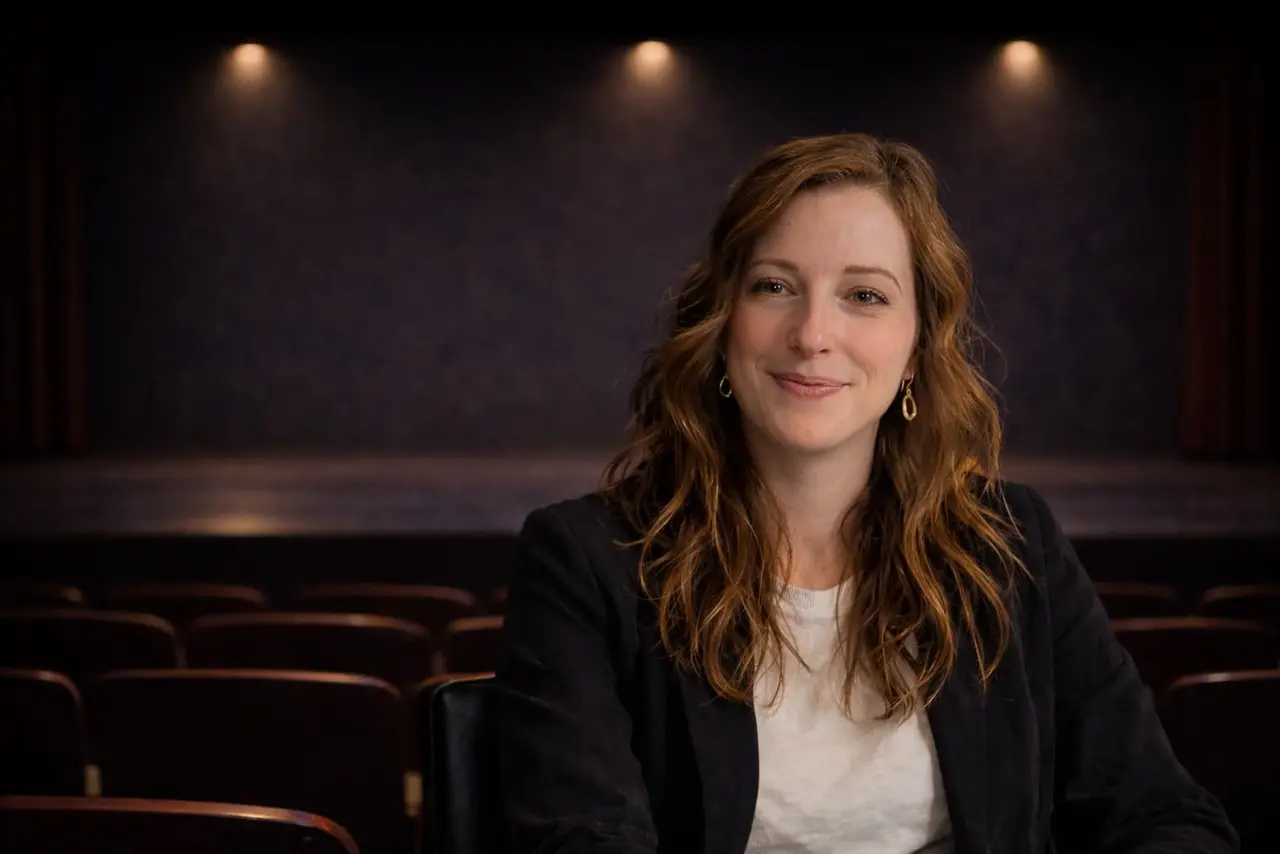 A woman with long wavy hair sits in an empty theater, facing the camera and smiling, with a dimly lit stage in the background.