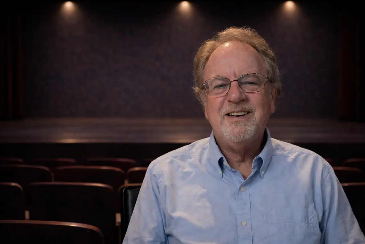 An older man with glasses and a light blue shirt sits smiling in an empty theater with rows of red seats and stage lights in the background.
