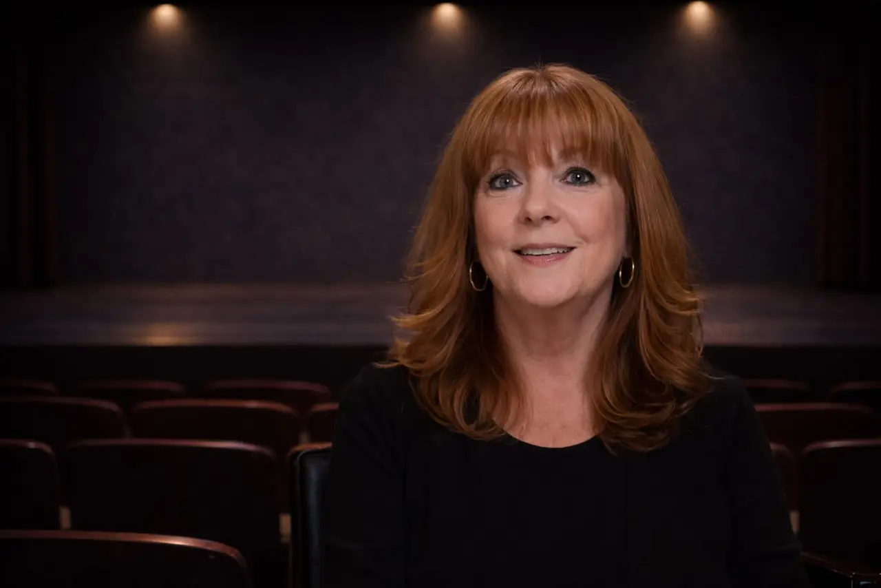 A woman with red hair and a black top sits in an empty theater, smiling at the camera. The stage and rows of seats are visible in the background.