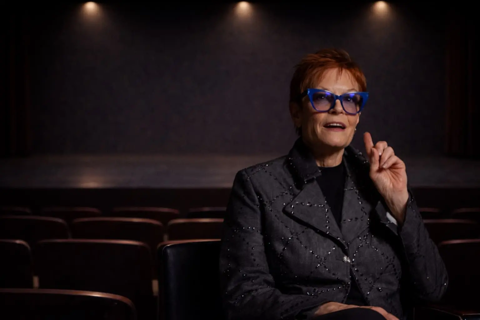A woman with short red hair and blue glasses sits in an empty theater, gesturing with her right hand while speaking.