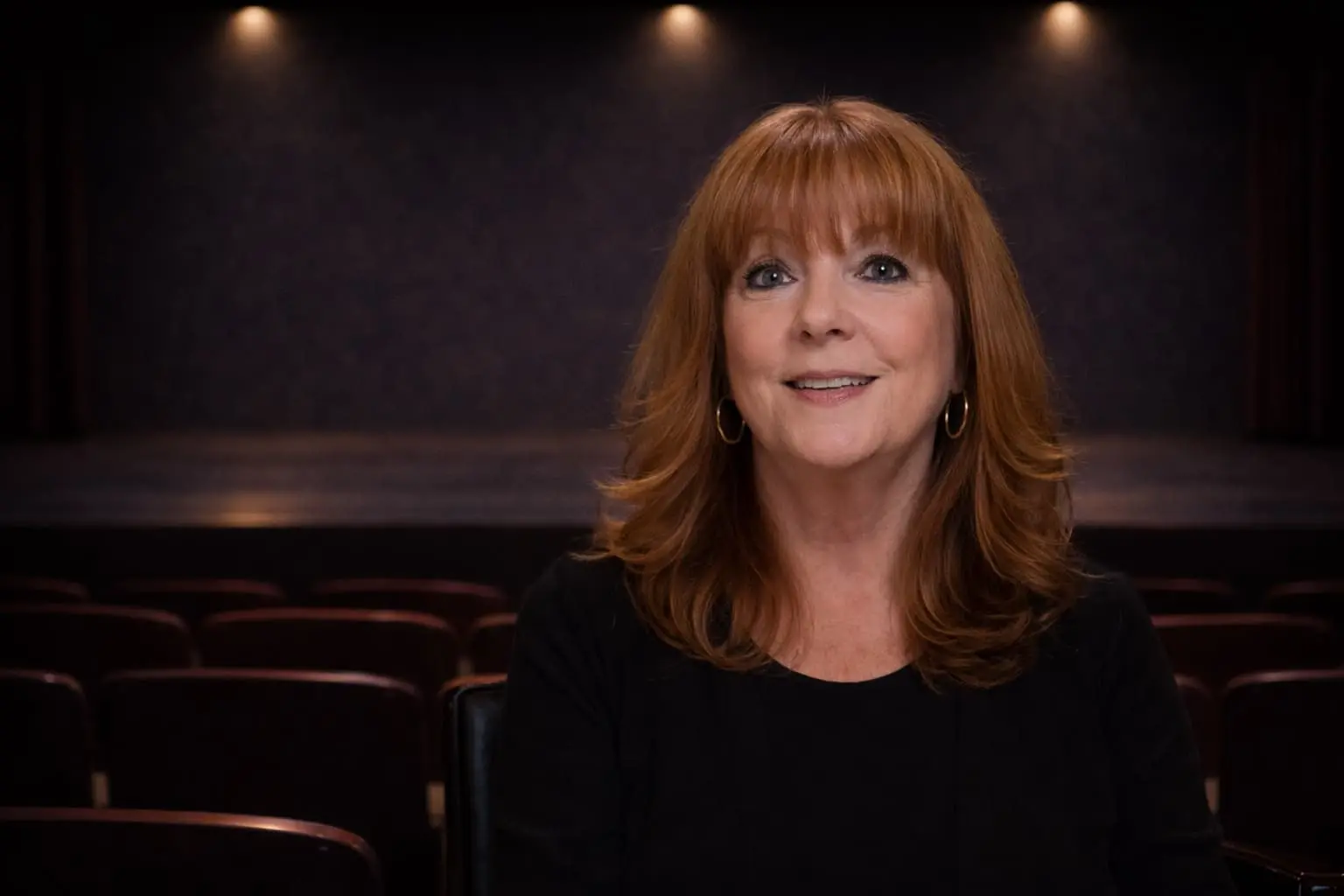 A woman with red hair and a black top sits in an empty theater, smiling at the camera. The stage and rows of seats are visible in the background.