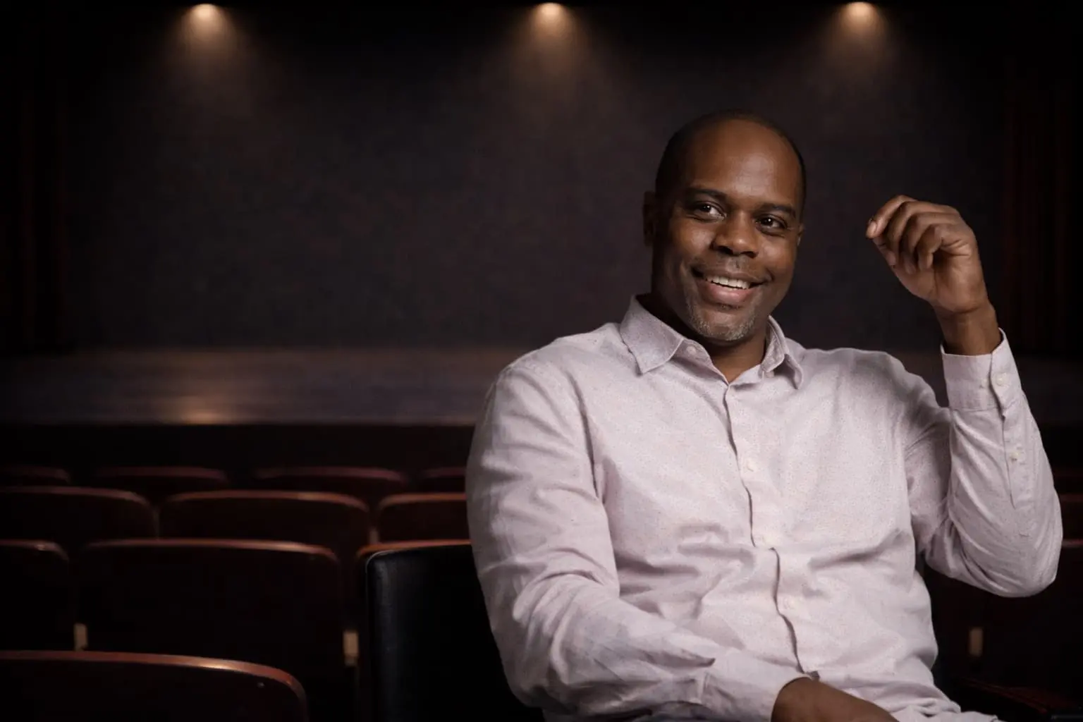A man in a light shirt sits and smiles in the foreground of an empty theater with rows of seats and a dimly lit stage behind him.