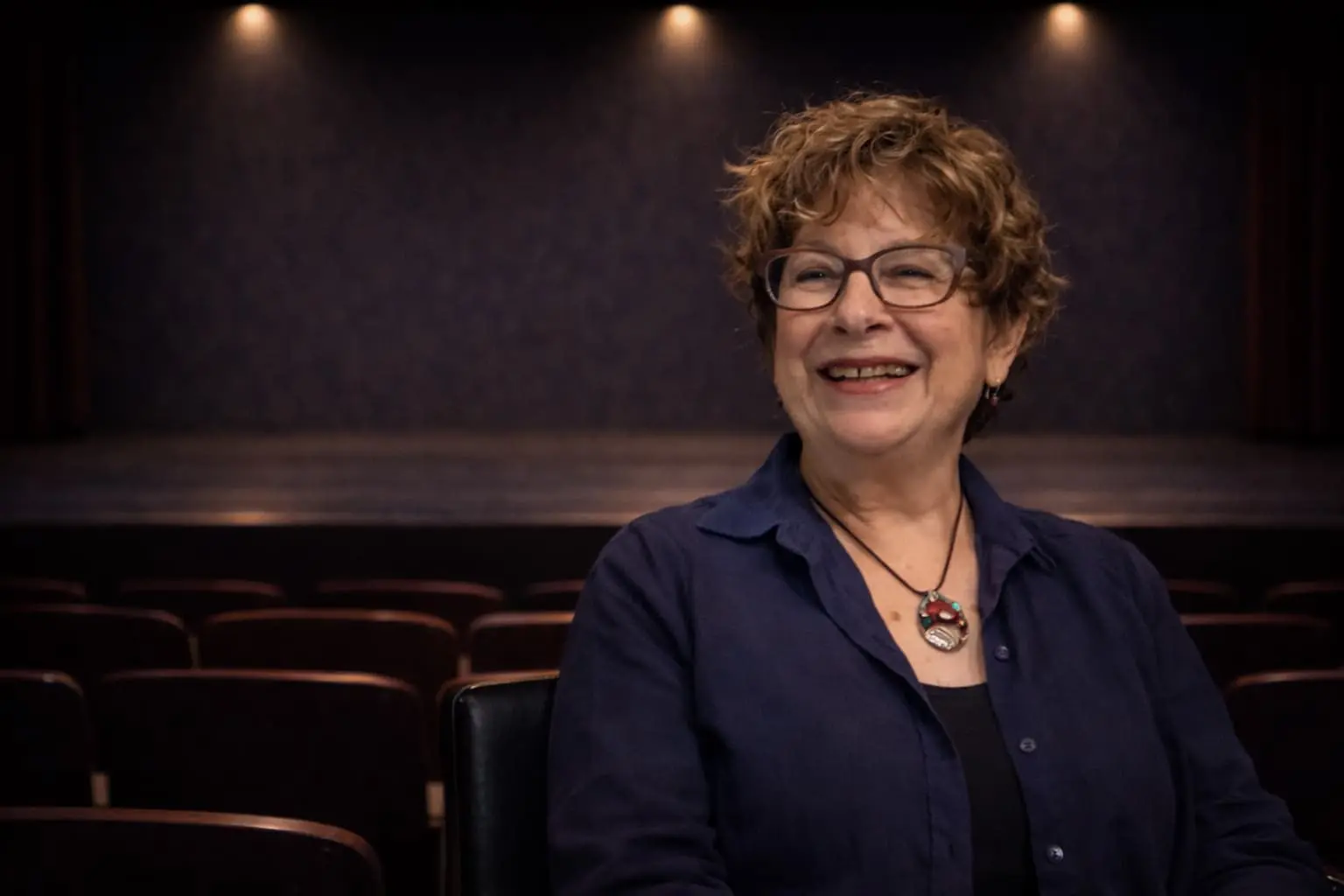 Smiling woman with curly hair and glasses sits in an empty theater with rows of seats and a dimly lit stage in the background.