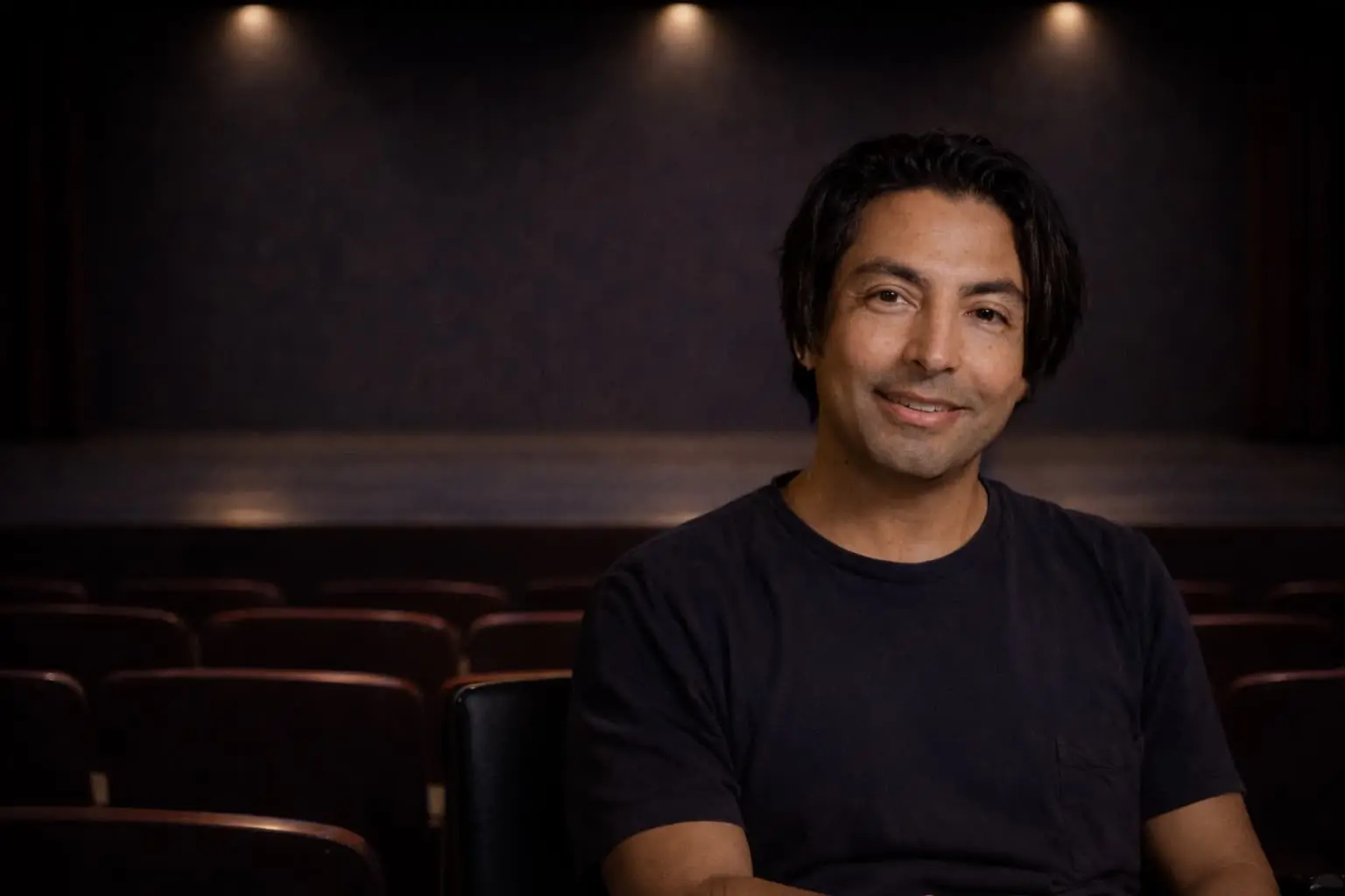 A man with medium-length dark hair and a black t-shirt sits smiling in an empty theater, with rows of seats and a dimly lit stage in the background.